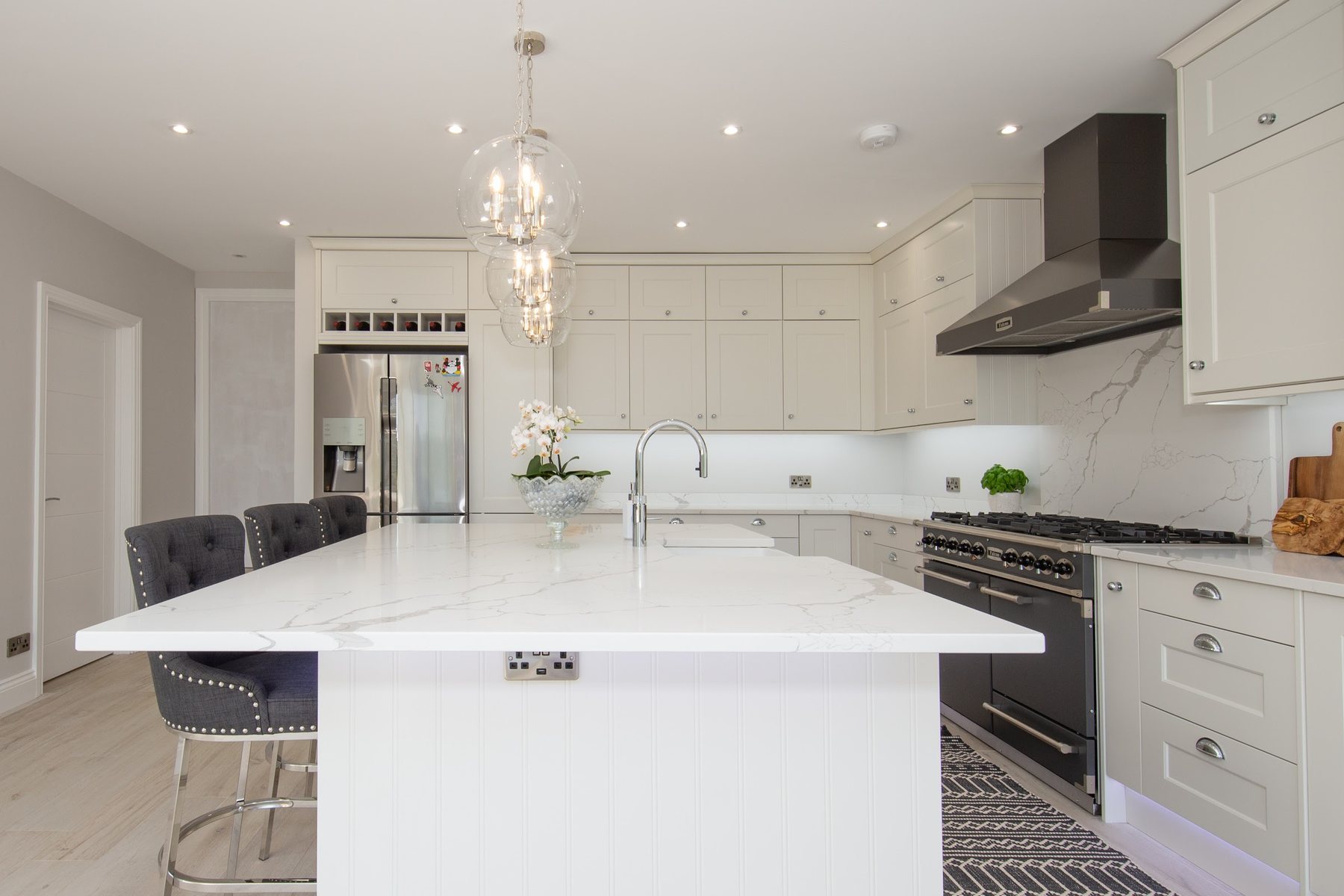 A large white shaker kitchen with a marble-effect quartz island, glass globe pendants and a black range cooker