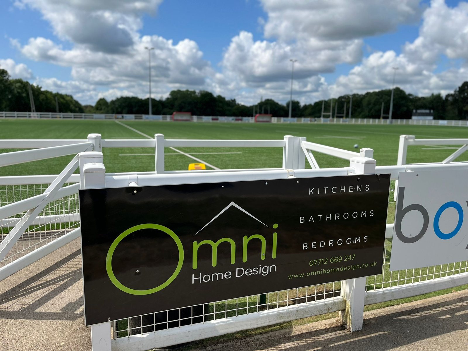 An Omni Home Design advertising board fixed to the railings beside a rugby pitch on a sunny day